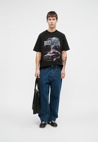 Mladic u crnoj majici s grafikom automobila, plavim širokim farmerkama, crnim tenisicama, drži crnu torbu za nošenje, stoji naslonjen na jednostavnu bijelu pozadinu.