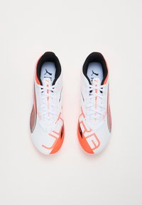 Hvite sportssko med teksturert overdel, svarte og oransje detaljer, samt et fremtredende logodesign på sidene og tungen.