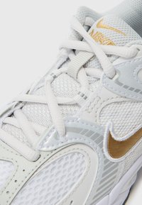 Biała sneakersy wykonane z siateczkowych i syntetycznych materiałów, z płaskimi sznurowadłami, metalicznymi złotymi akcentami oraz teksturowaną podeszwą zapewniającą przyczepność.