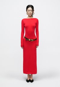 Czerwona sukienka z długim rękawem o dopasowanym kroju, z teksturowanej tkaniny, z brązowym paskiem z złotą klamrą. Czarne buty z noskiem w szpic.