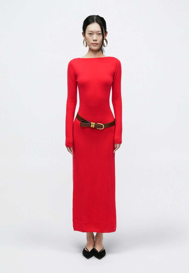 Czerwona sukienka z długim rękawem o dopasowanym kroju, z teksturowanej tkaniny, z brązowym paskiem z złotą klamrą. Czarne buty z noskiem w szpic.