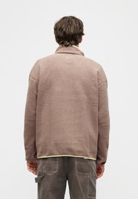 Άνδρας που φοράει ανοιχτό καφέ φλις μπουφάν και σκούρο παντελόνι cargo, να στέκεται με την πλάτη προς το μέρος μας πάνω σε απλό ανοιχτό φόντο.