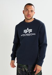 Sweatshirt à col rond bleu marine en tissu doux, avec un logo blanc et le texte "ALPHA INDUSTRIES" avec une finition texturée.