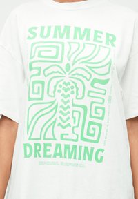 Λευκό βαμβακερό t-shirt με μεγάλο πράσινο γραφικό που απεικονίζει μια στυλιζαρισμένη φιγούρα και γεωμετρικά μοτίβα. Το κείμενο αναφέρει "SUMMER DREAMING" και το όνομα της μάρκας.