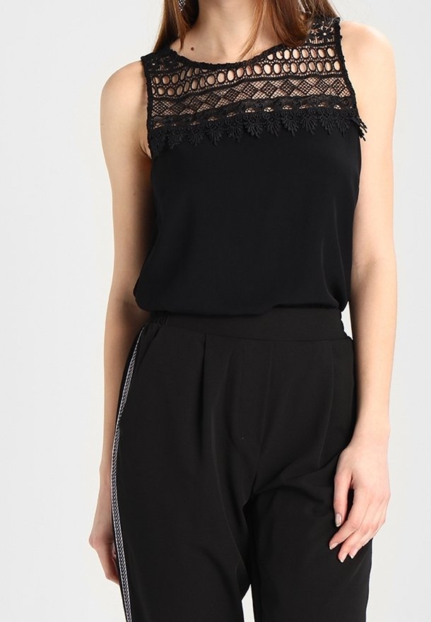 Haut noir sans manches avec empiècement en dentelle et bordure florale, porté avec un pantalon noir taille haute à rayure blanche sur le côté.