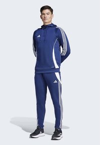 Marinblå träningsdräkt med vita detaljer och tre vita ränder på ärmar och byxor. Huva på tröjan, elastisk midjeresår och sportskor.