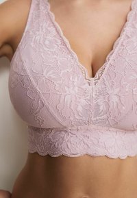 Bralette in pizzo di un tenue rosa con motivo floreale, design a V profonda, bordi ondulati e tessuto testurizzato, per un aspetto delicato.