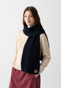 Fular tricotat de culoare bleumarin, așezat peste un pulover bej deschis, cu o textură cu nervuri și un detaliu în formă de logo circular la capătul fularului.