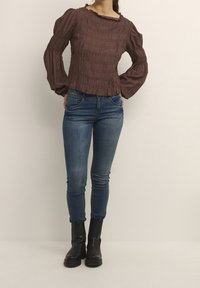 Blusa marrone con arricciature e maniche a palloncino, abbinata a jeans skinny blu e stivaletti neri. Tessuto morbido con una texture elastica.