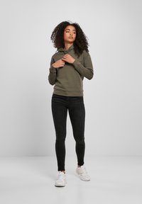 Donna con i capelli ricci che indossa un felpa oliva, jeans skinny neri e sneakers bianche, in piedi su uno sfondo grigio chiaro uniforme.