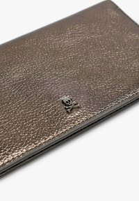 Cartera de bronce metálico con superficie texturizada; presenta un pequeño adorno de calavera y huesos cruzados en negro. Forma rectangular compacta.