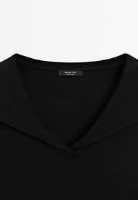 Pull en maille noire avec un large col rabattu et une petite étiquette portant la mention "Massimo Dutti Made in China" au niveau de l'encolure.