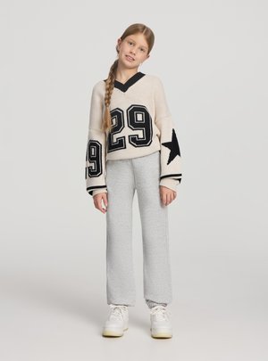 Ragazza giovane con lunga treccia che indossa un maglione beige con il numero 29 e disegni di stelle, pantaloni della tuta grigi e scarpe da ginnastica bianche, in piedi su sfondo semplice.