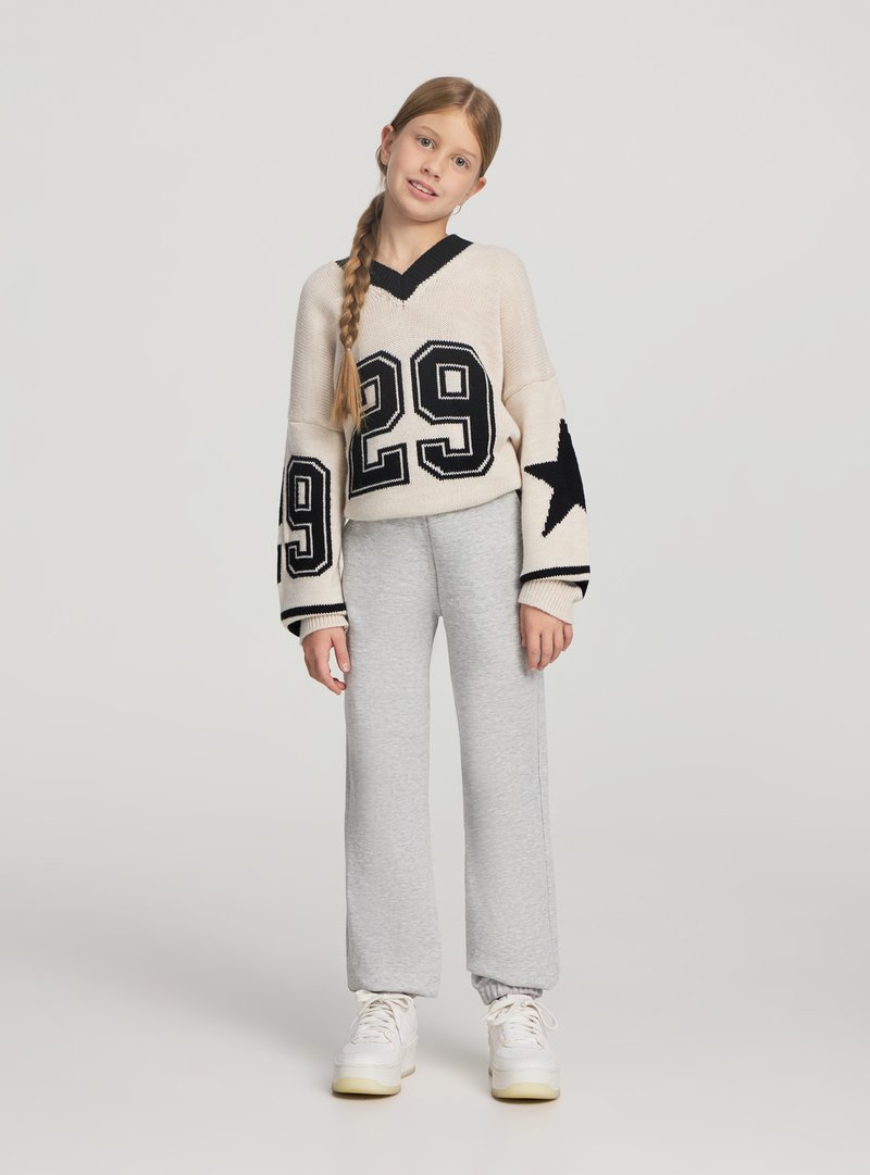 Ragazza giovane con lunga treccia che indossa un maglione beige con il numero 29 e disegni di stelle, pantaloni della tuta grigi e scarpe da ginnastica bianche, in piedi su sfondo semplice.