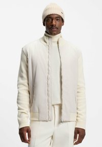 Lys beige bomberjakke med lynlås foran, ribbede manchetter og høj krave, parret med en cremet turtleneck og matchende underdele.