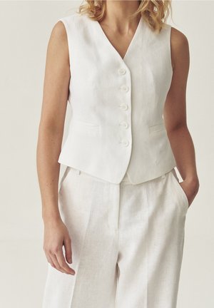 Femme portant un gilet blanc sans manches boutonné et un pantalon blanc taille haute assorti, une main dans la poche, sur un fond uni.