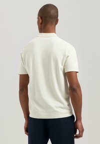 Lys beige polo skjorte med korte ermer og ribbestrikket krave. Glatt tekstur, tettsittende design, vist fra baksiden mot en nøytral bakgrunn.