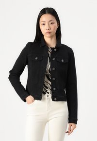 Svart denimjakke med knapper, to frontlommer og en strukturert krage. Modellen kombinerer den med et mønstrete plagg og lyse bukser.