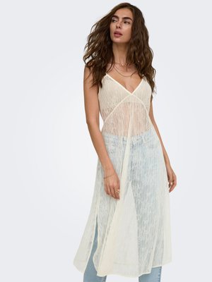 Femme portant une robe sans manches en dentelle crème transparente sur un jean bleu clair, avec des cheveux ondulés lâches et des bijoux discrets sur un fond uni.