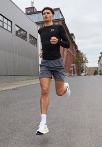 Atleta che corre su una strada, indossando una maglietta nera attillata a maniche lunghe e pantaloni corti grigi, con scarpe da corsa bianche e calzini alla caviglia.