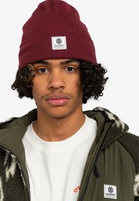Giovane con capelli ricci che indossa un berretto bordeaux e una giacca in pile verde con t-shirt bianca, che guarda direttamente davanti a sé.