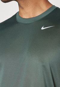 Ciemnozielona koszulka sportowa Nike z okrągłym dekoltem, krótkimi rękawami i małym białym logo swoosh na górnej, lewej części klatki piersiowej.