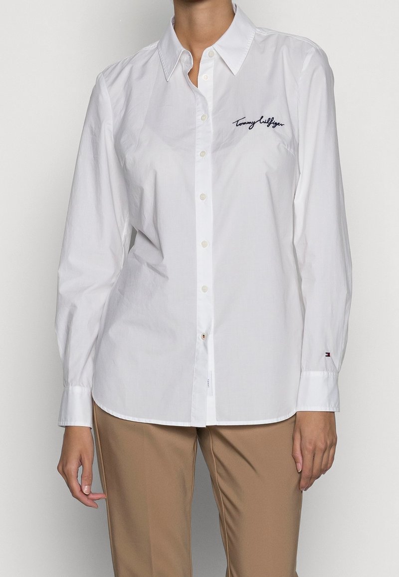 Femme portant une chemise blanche à manches longues boutonnée et un pantalon beige, la chemise porte le logo "Tommy Hilfiger" brodé sur la poitrine.