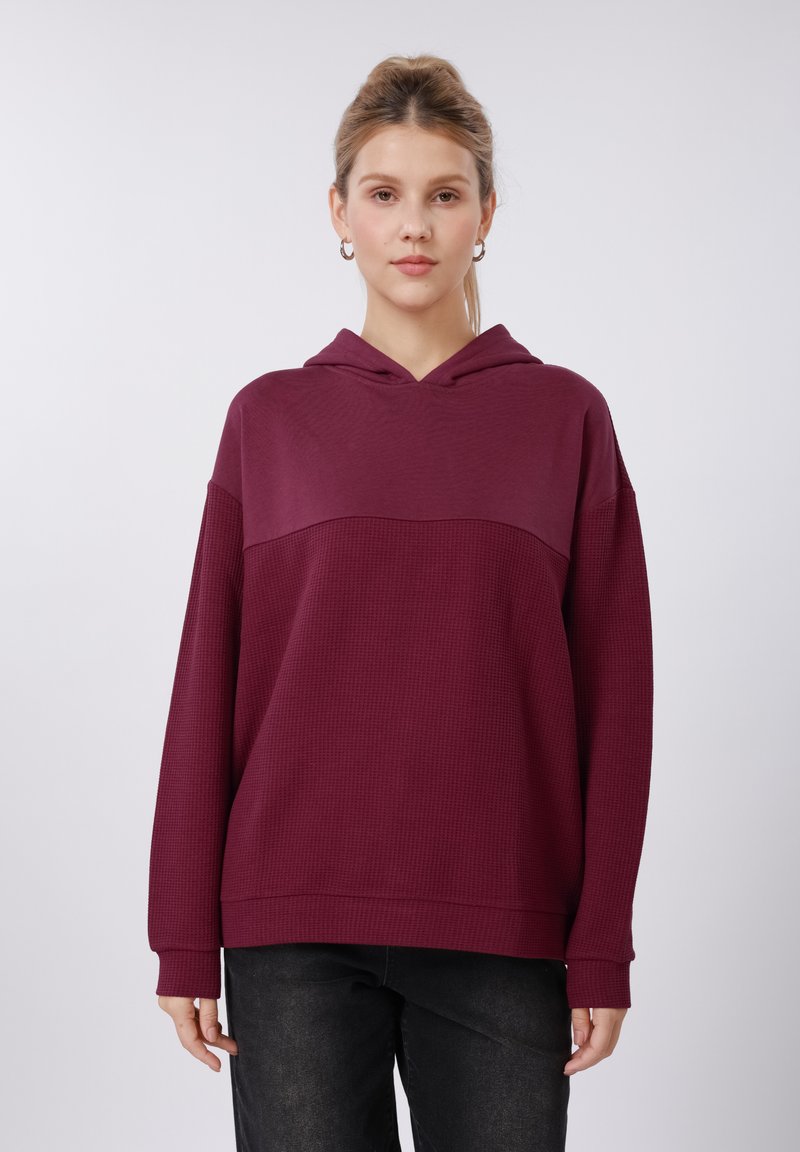 Sweat-shirt à capuche bordeaux avec partie supérieure texturée ; présente un tissu côtelé et des manches longues. Associé à un jean foncé, mettant en avant un style décontracté.