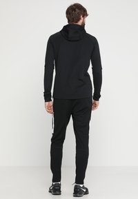 Schwarzer sportlicher Kapuzenpullover mit Kapuze und langen Ärmeln, kombiniert mit schwarzen Hosen, die weiße Seitestreifen aufweisen. Der Stoff wirkt glatt und dehnbar.