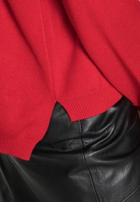 Czerwony sweter z dzianiny o prążkowanej teksturze i bocznych rozcięciach, w zestawieniu z gładką czarną skórzaną spódnicą. Skupienie na wyrazistym kontraście kolorów.