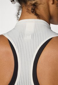 Top sportivo bianco e nero con collo alto, caratterizzato da una texture a costine e un logo sul retro. Contrasto di colore sulle aperture per le braccia e sul collo.