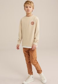 Chemise beige à manches longues texturée avec un emblème rond, associée à un pantalon cargo marron avec des poches latérales et des baskets montantes blanches.