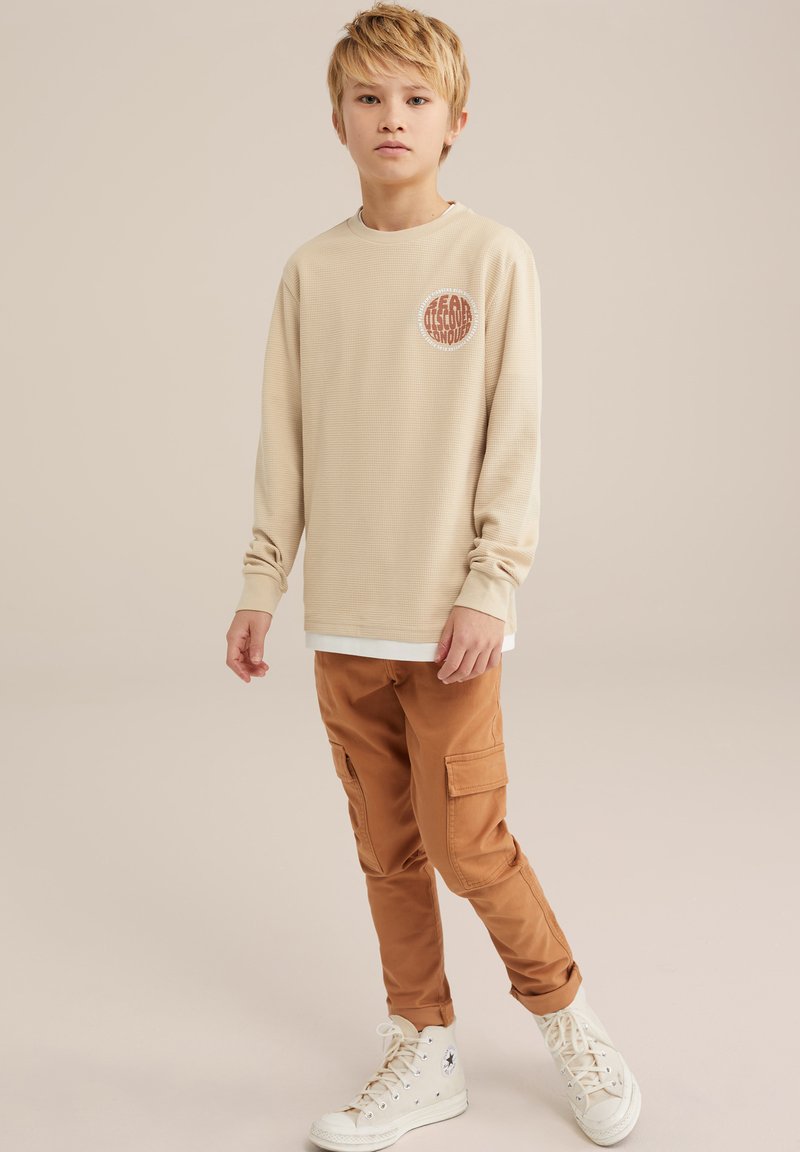 Chemise beige à manches longues texturée avec un emblème rond, associée à un pantalon cargo marron avec des poches latérales et des baskets montantes blanches.