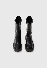 Bottes de cheville en cuir noir avec une texture lisse, bouts carrés et semelles en caoutchouc robustes. Équipées de languettes pour un enfilage facile.