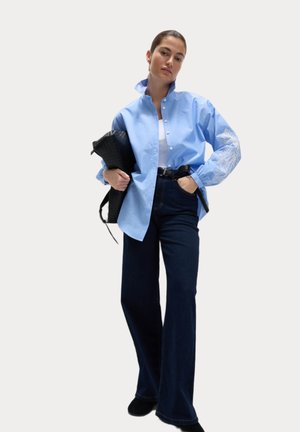 Camicia azzurra chiara con ricami bianchi intricati sulle maniche, abbinata a jeans a vita alta blu scuro svasati e una borsa nera con texture.