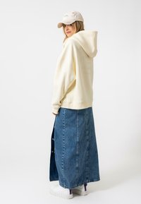 Sudadera crema con capucha, combinada con una falda larga de mezclilla. Zapatillas blancas con detalles púrpuras. Diseño casual y de corte relajado.