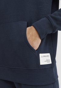 Sudadera navy con un bolsillo tipo canguro y puños acanalados. Incluye una etiqueta blanca con los detalles de la marca cosida a un lado. Tejido suave y texturizado.
