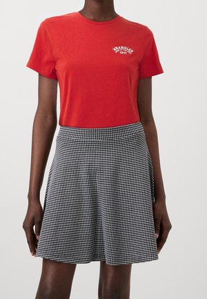 Červené bavlněné tričko s vyšívaným logem "Wrangler" na hrudi, v kombinaci s černo-bílou kostkovanou sukní s vysokým pasem, sahající ke kolenům.
