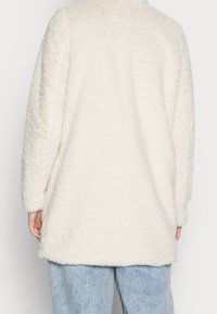 Prenda exterior de piel sintética en color crema, con un corte relajado, mangas largas y un dobladillo ligeramente redondeado. La textura parece suave y esponjosa.