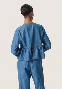 Denim peplumblus med långa ärmar, samlad midja och rundad nederkant. Ljust blått tyg med en subtil textur, försedd med rundad halsringning.