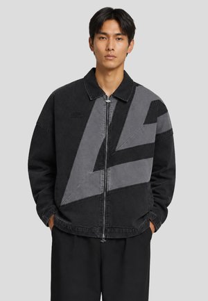 Uomo che indossa una giacca nera e grigia con motivo geometrico con zip e colletto, mani nelle tasche, in piedi davanti a uno sfondo grigio chiaro uniforme.