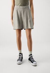 Lysgrå, plisserede shorts med en glat tekstur, parret med sorte høje sneakers og lyse sokker.