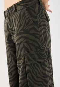 Cargo pants made of a textured fabric featuring a green and black zebra-stripe pattern. Includes a side pocket and metal button closure.