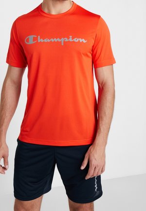 Hombre con camiseta Champion naranja brillante y pantalones cortos deportivos negros, de pie frente a un fondo claro y liso.