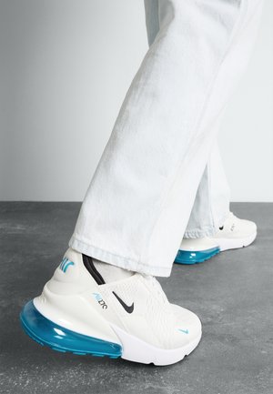 Personne portant des baskets blanches Nike Air Max 270 avec un coussin d'air bleu, debout sur un sol gris, portant un jean bleu clair et des chaussettes.