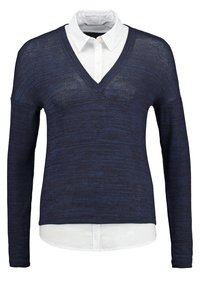 Granatowy sweter z dekoltem w serek nałożony na białą koszulę z kołnierzykiem i długimi rękawami.