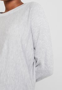 Maglione grigio chiaro realizzato in tessuto morbido, con un ampio scollo e maniche leggermente oversize. La texture appare liscia con lievi variazioni.