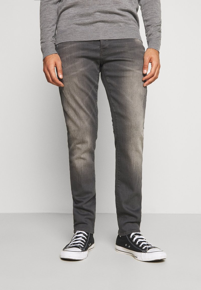 Vaqueros de mezclilla grises con acabado desgastado, corte slim, diseño de cinco bolsillos y zapatillas negras bajas con suelas y cordones blancos.