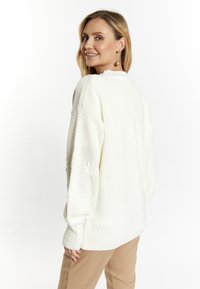Cremfarbener, gestrickter Pullover mit strukturierten Ärmeln und gerippten Bündchen. Mit Rundhalsausschnitt und lässiger Passform, getragen zu beigen Hosen.
