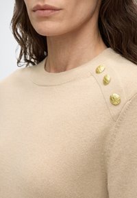 Femme portant un pull en maille beige avec un col rond et trois boutons dorés texturés sur l'épaule gauche.